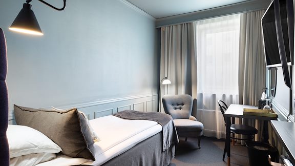 Ett litet hotellrum med en liten säng, en fåtölj, ett skrivbord och en tv.