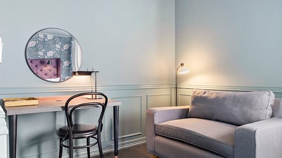 Ett skrivbord med en stol. En spegel hänger på väggen. Bredvid står en liten soffa.