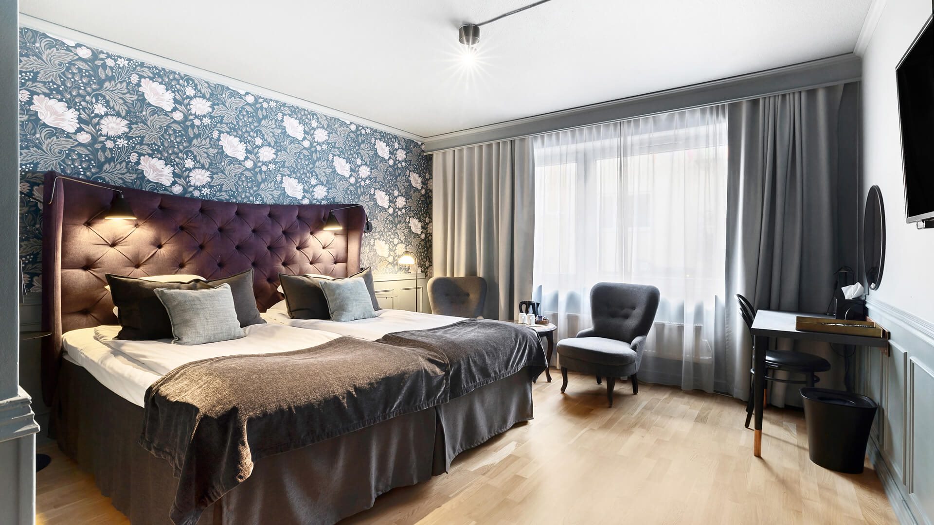 Hotellrum med en stor dubbelsäng. Bredvid sängen finns två fåtöljer och ett skrivbord. På väggen hänger en tv.