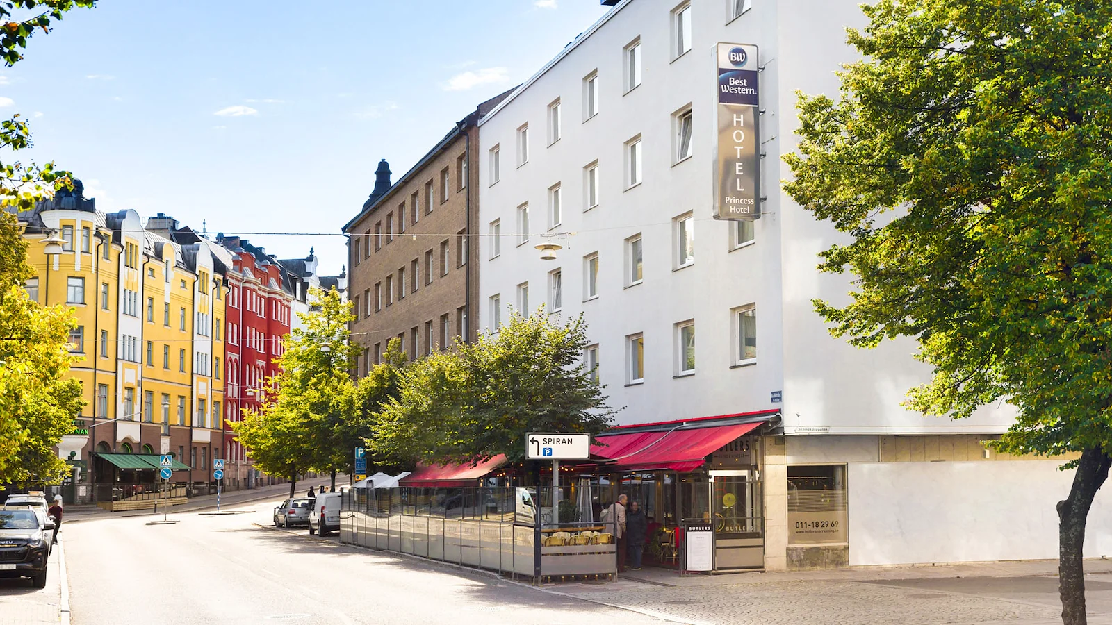 Fasadbild på Princess Hotel i Norrköping. Ett vitt, äldre hus med andra färggalda hus runtom kring vid en stadsväg.